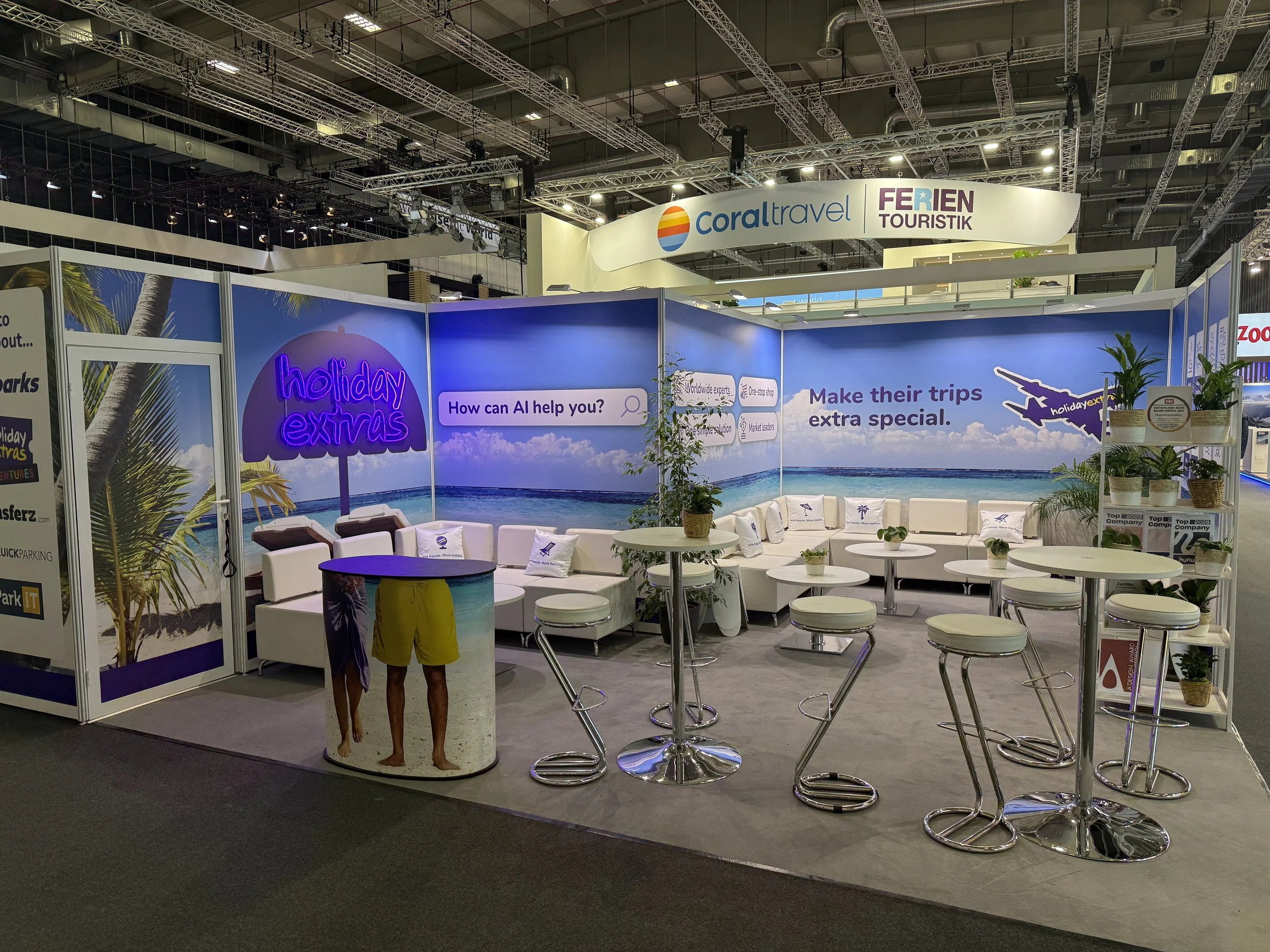 Messestand mit einem Strandmotiv, weißen Sitzmöbeln, Tischen und Pflanzen, Werbung für Reise-Extras, Slogans wie 'Make their trips extra special', und dem Claim "Less hassle. More holiday."