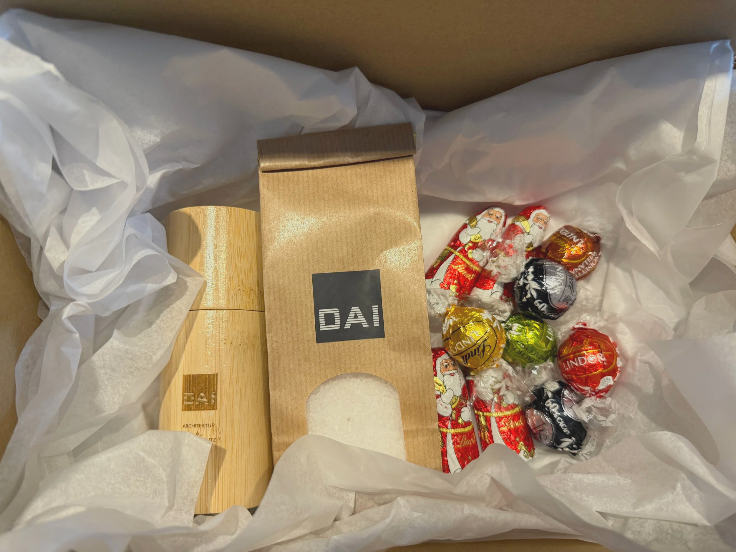 Geschenkbox mit einer Holzkiste, einer braunen Papiertüte mit DAI-Aufdruck, bunten Weihnachtsschokokugeln und weihnachtlichem Geschenkband, eingepackt in weißem Papier