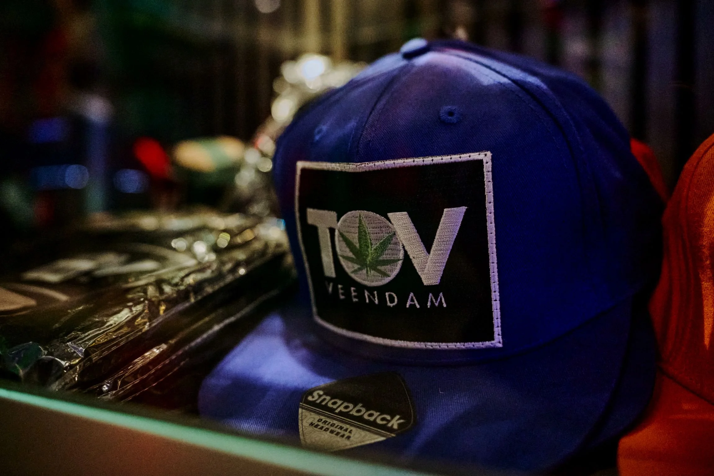 Blauwe pet met ontwerp met de tekst 'TOV VEENDAM' en een cannabisplant, liggend op een tafel in een donkere omgeving.