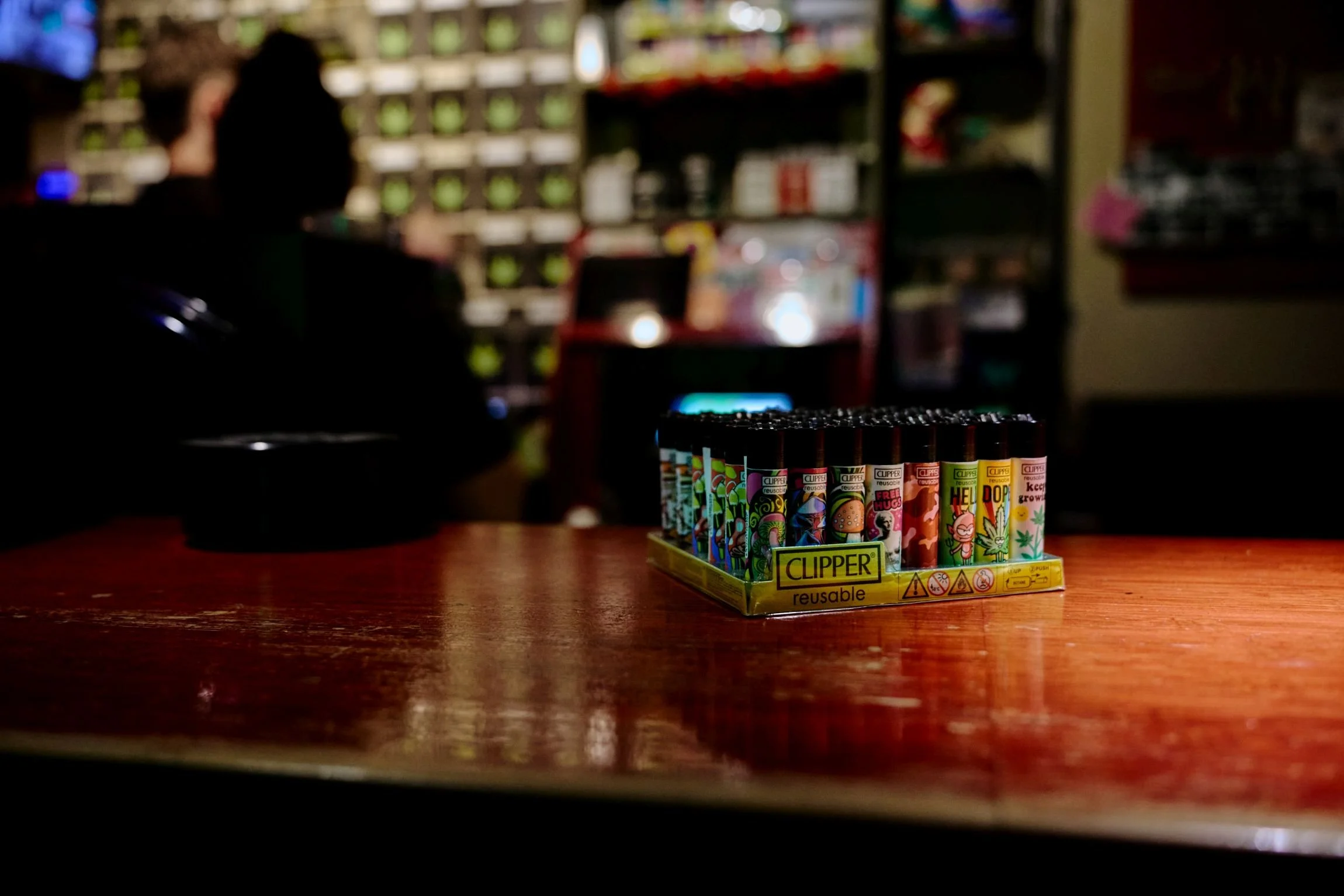 Rijen vape penne op een houten tafel, met een achtergrond van een gestructureerde muur en mensen die in een winkel of café staan.