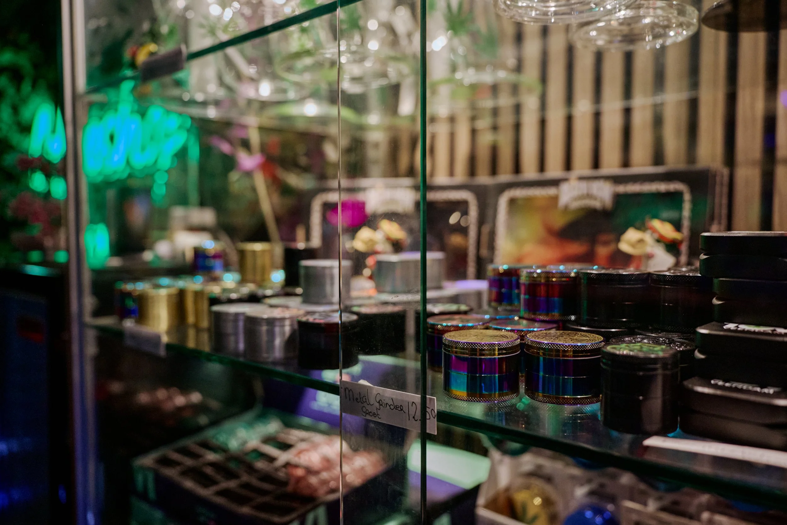 Een glazen displaykast met metalen grinder pots en andere rookaccessoires, met een neonbord en houten achtergrond.