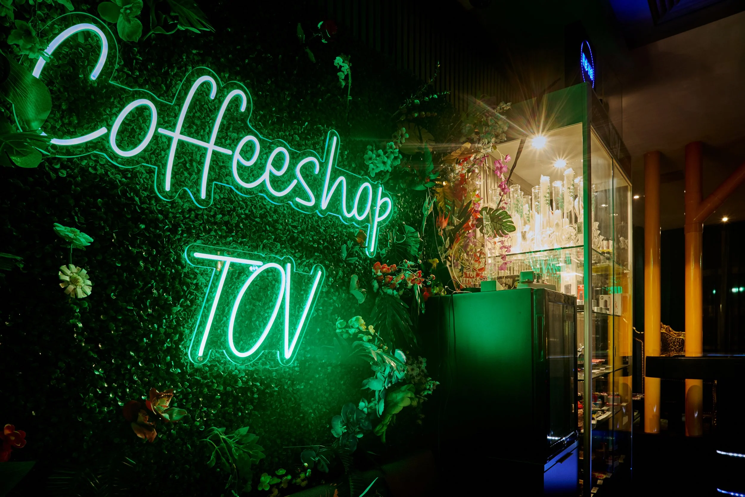 Groene neonletters spelling 'CoffeeShop TOV' hangen aan een groene muur met planten, naast een glazen displaykast gevuld met bloemen en decoraties, binnen een winkel of café.