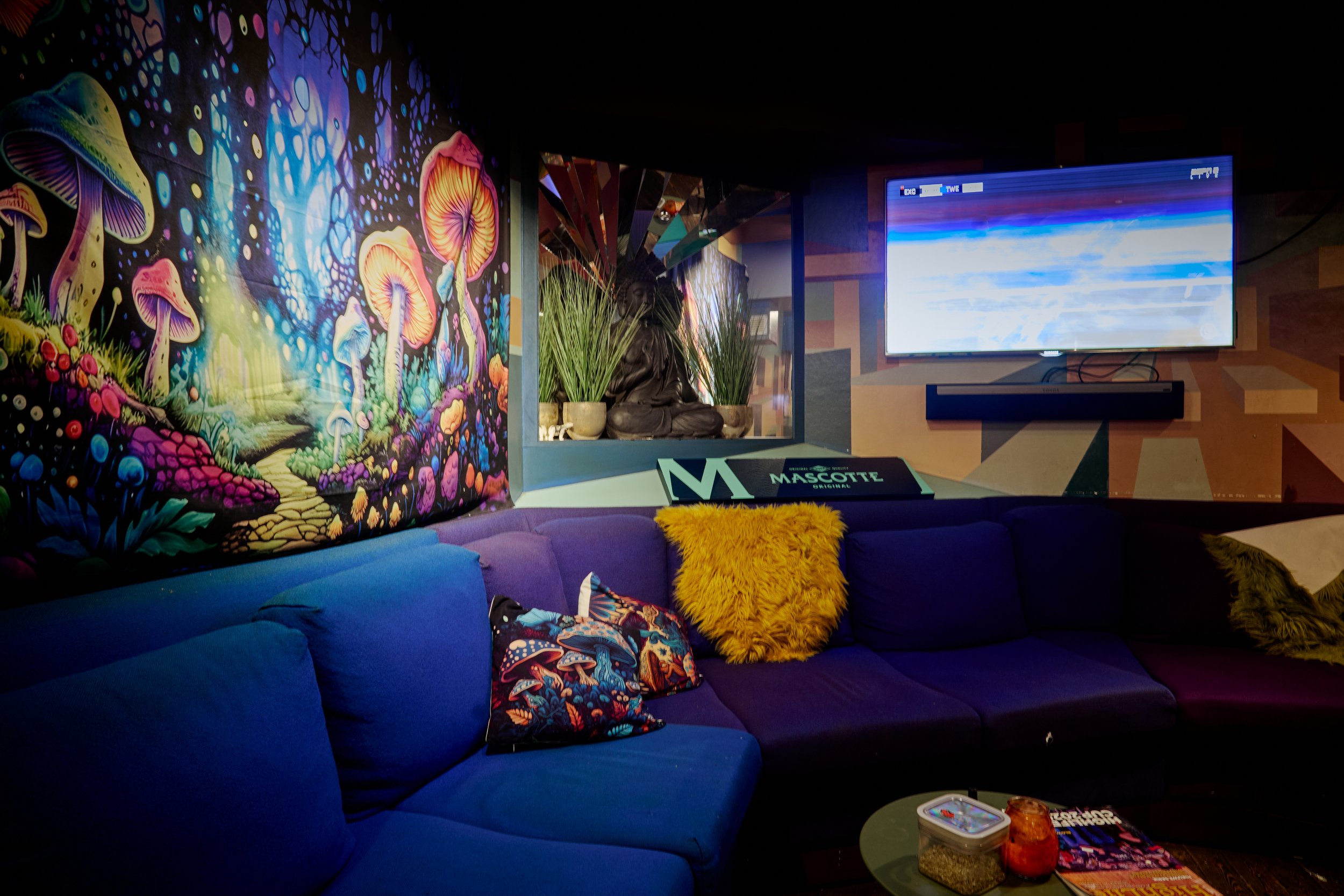 Knusse woonkamer met een kleurrijk muurschildering van paddenstoelen, een blauwe bank met verschillende kussens, een tafel met snacks en tijdschriften, een tv-scherm aan de muur, en decoratieve planten.