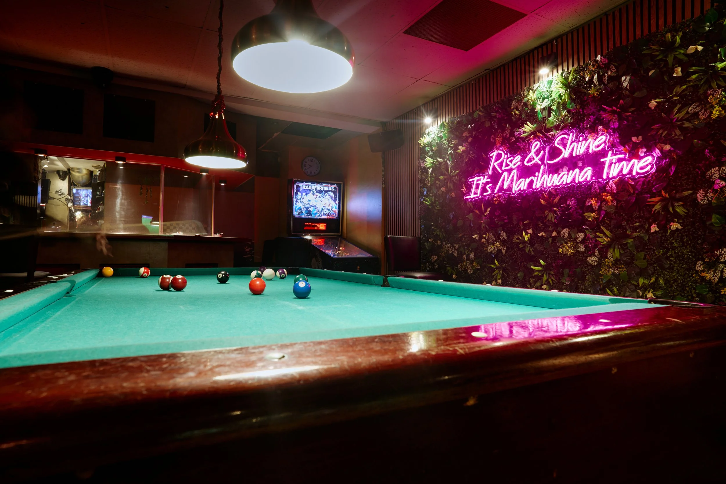 Interieur van een bar met een biljarttafel, neonbord met de tekst 'Rise & Shine It's Marihuana Time', een muur met bloemen, een arcade-spel en hanglampen.