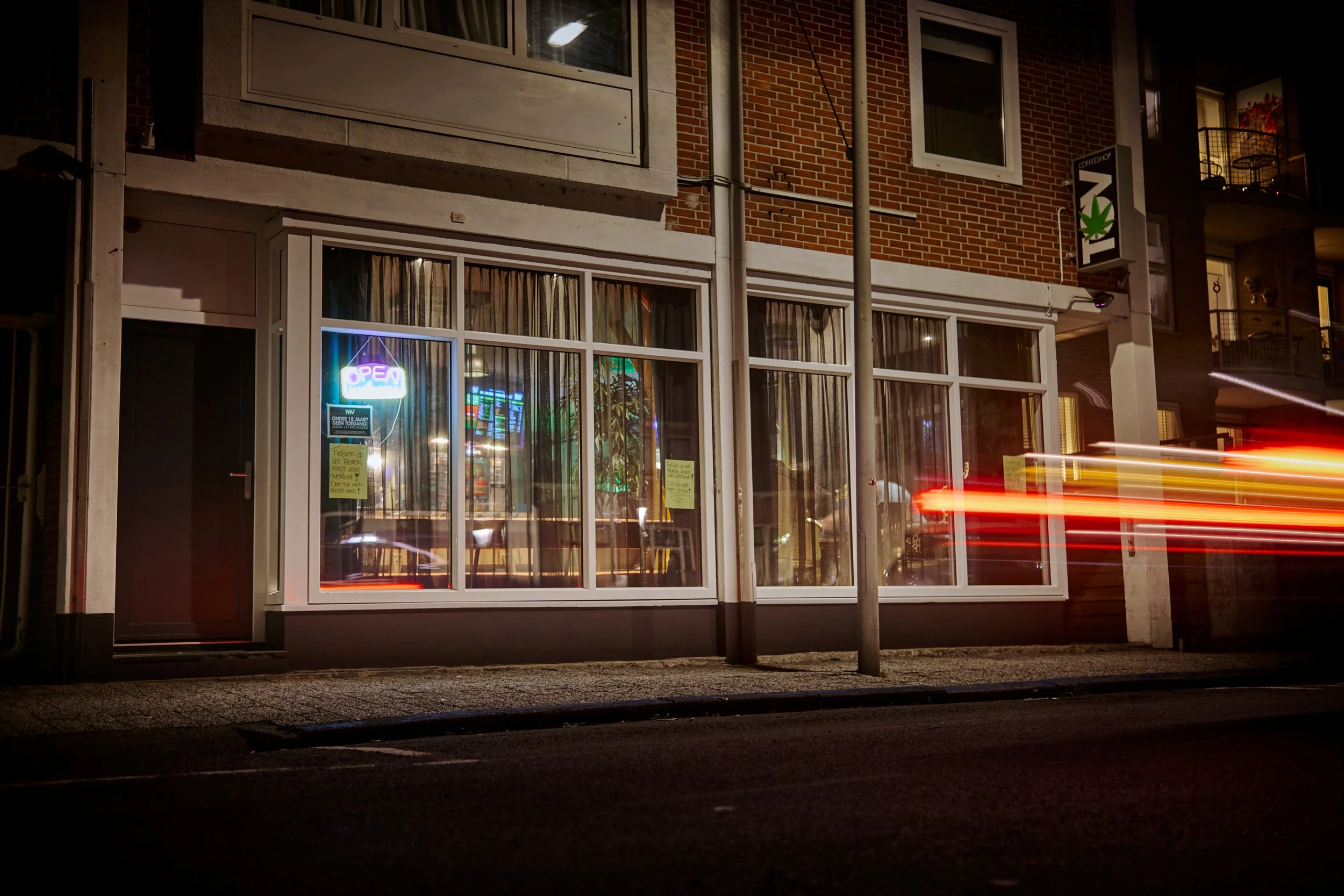 Foto van een nachtzicht van een winkel met grote ramen, een neon 'OPEN'-bord en binnenverlichting, met een straat en auto’s met lichtsporen voor de winkel.
