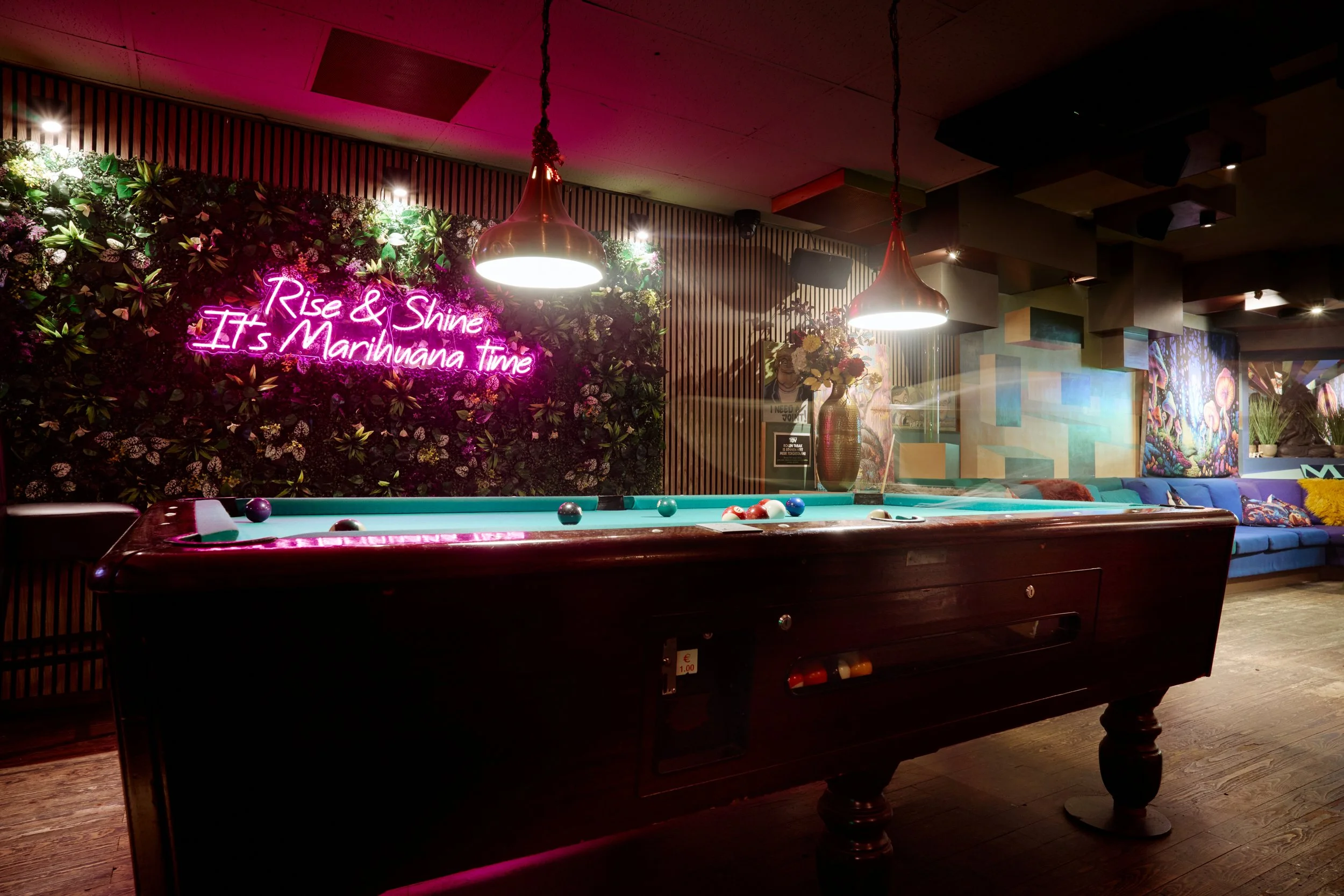 Een biljarttafel in een trendy bar of lounge, verlicht door hanglampen, met een kleurrijke muur en neonbord met tekst 'Rise & Shine It's Marijuana Time' op de achtergrond, versierd met bloemen en planten.