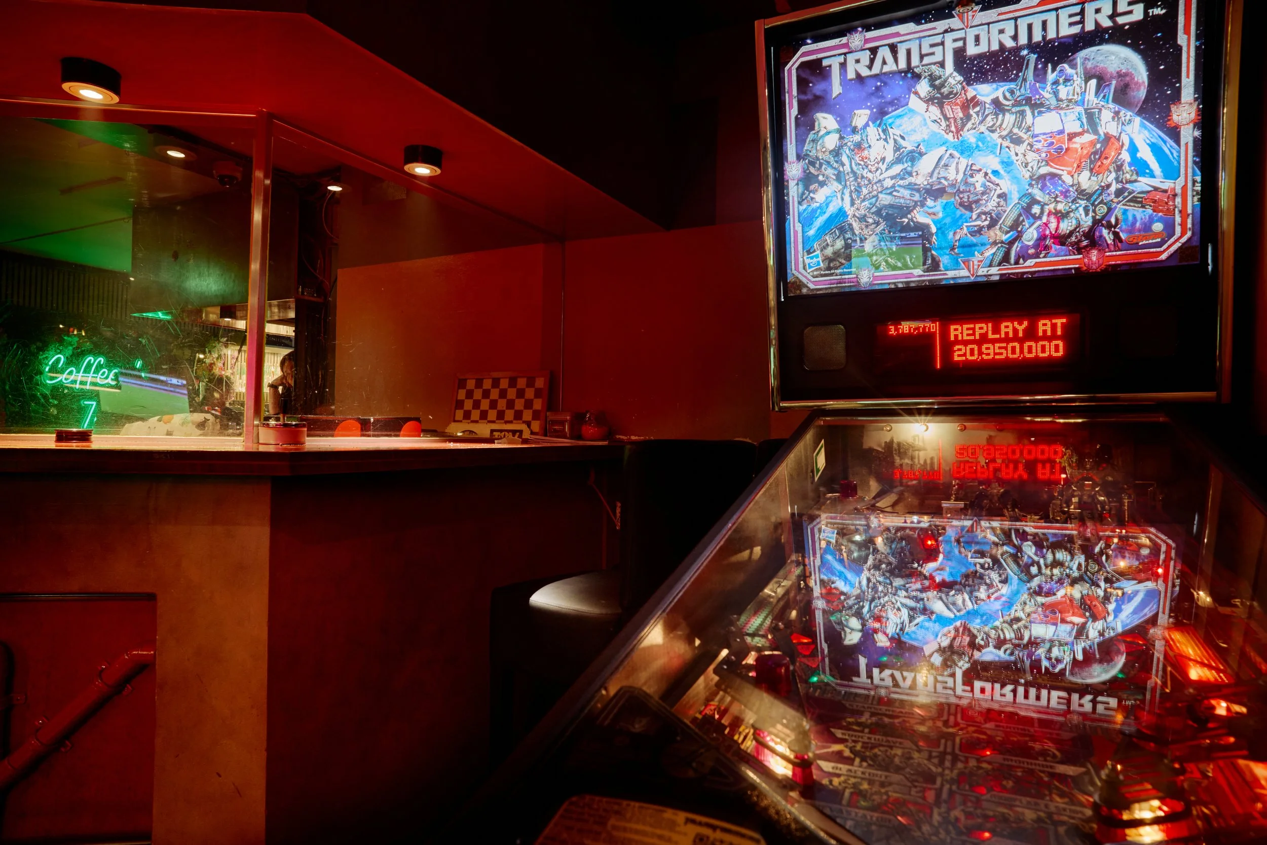 Pinball machine met Transformers afbeelding, weergegeven in een bar met rood gekleurde muren en een neon 'Coffee' sign.