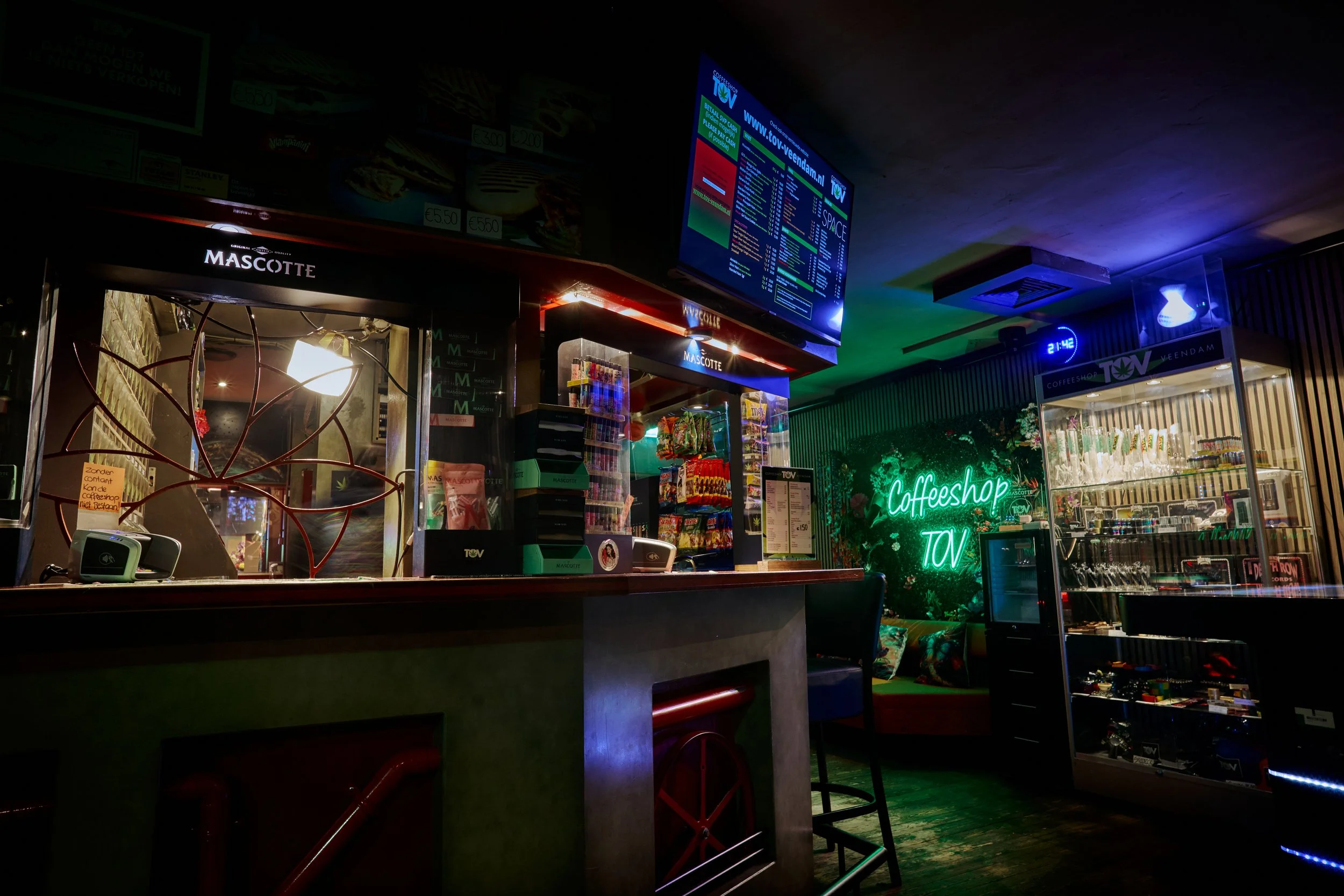 Interieur van een café of eetcafé met een menukaart, neonverlichting met de tekst 'Coffeeshop TOV', een balie met kassasystemen, en een displaykast met snacks en drankjes.