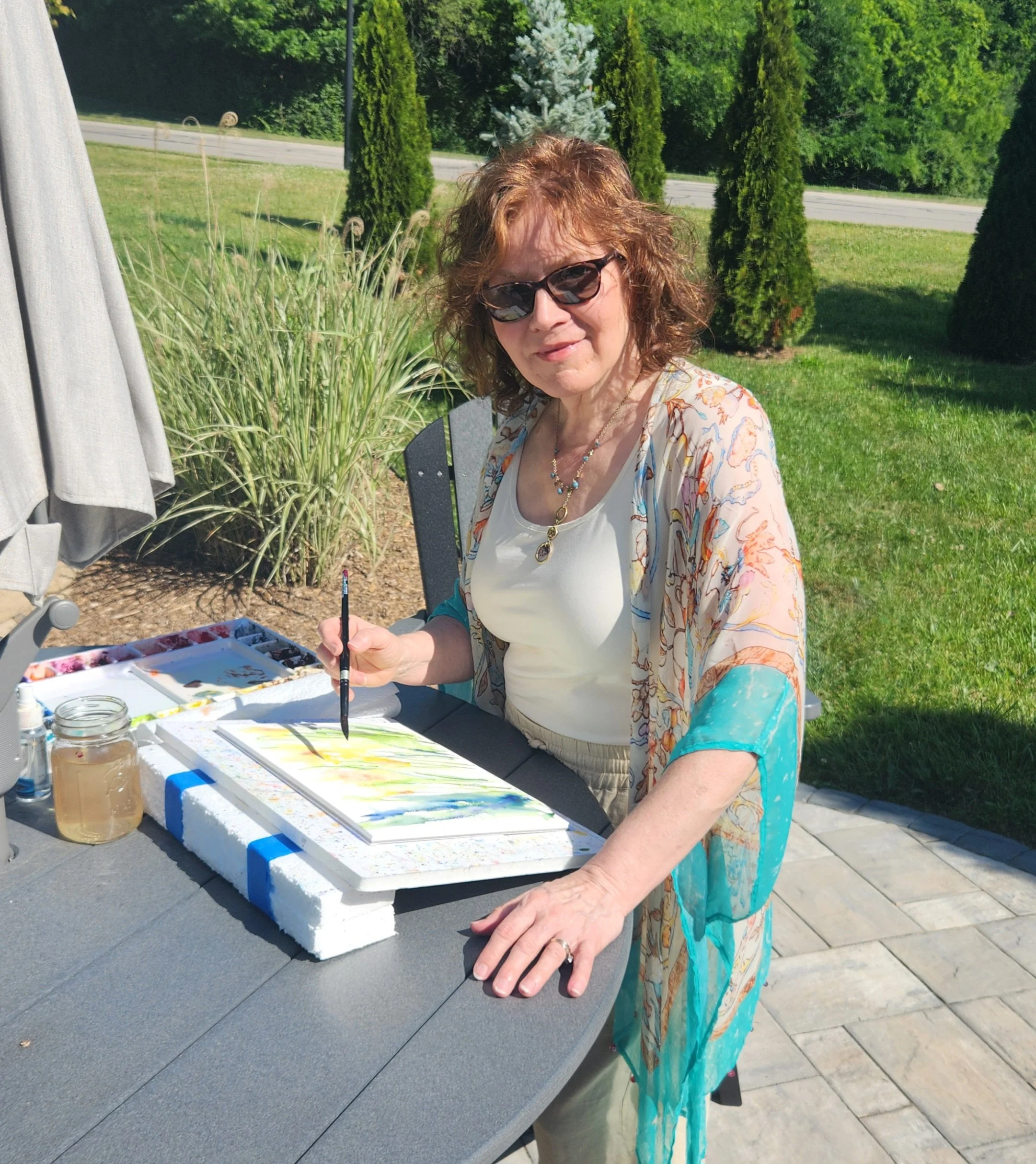A woman with curly red hair and sunglasses painting a landscape on watercolor paper outdoors, sitting at a table with art supplies.