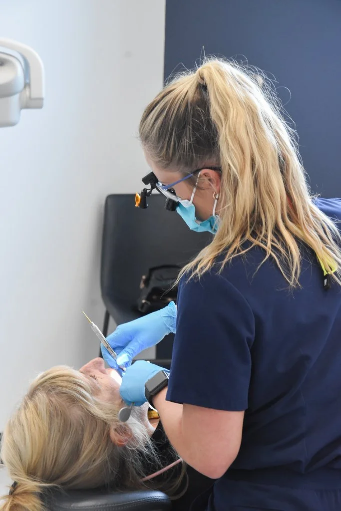 A dental professional wearing protective gear, including gloves, mask, and magnifying glasses, administering anesthesia to an elderly patient in a dental clinic.