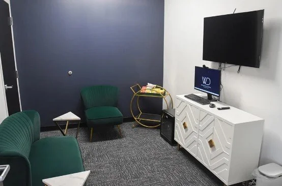 A small office or waiting room with a blue wall, green chairs, a white cabinet, a wall-mounted TV, and a computer monitor.