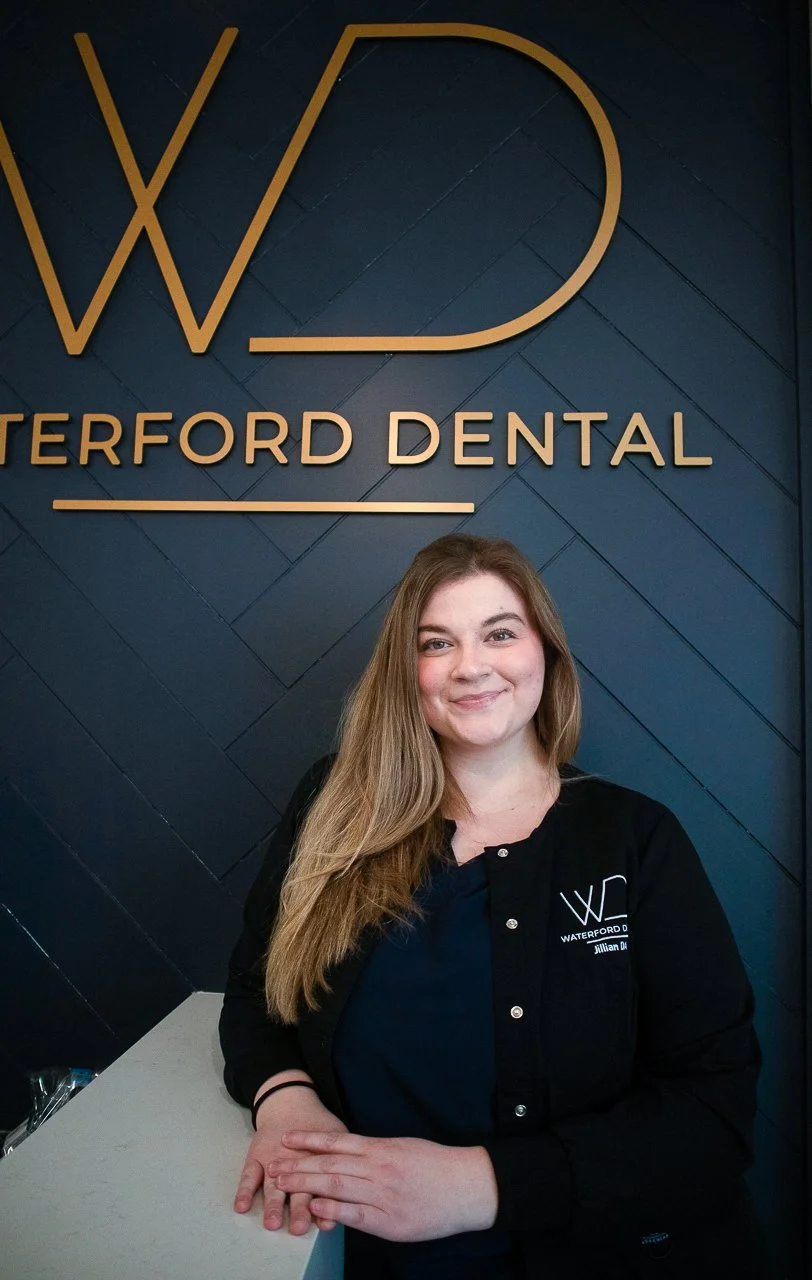 A woman with long, light brown hair smiling while standing in front of a sign that reads "Waterford Dental" on a dark blue wall, wearing a black jacket with a logo.