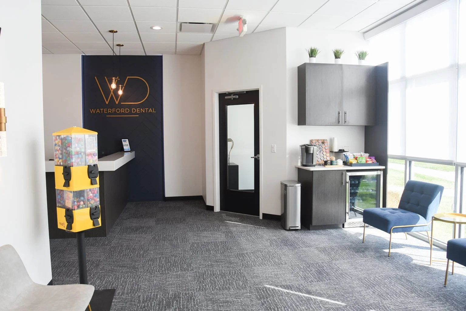 Waiting area of Waterford Dental office with a pop-up gumball machine, a coffee station with snacks, cups, utensils, a small refrigerator, and a blue lounge chair near large windows.