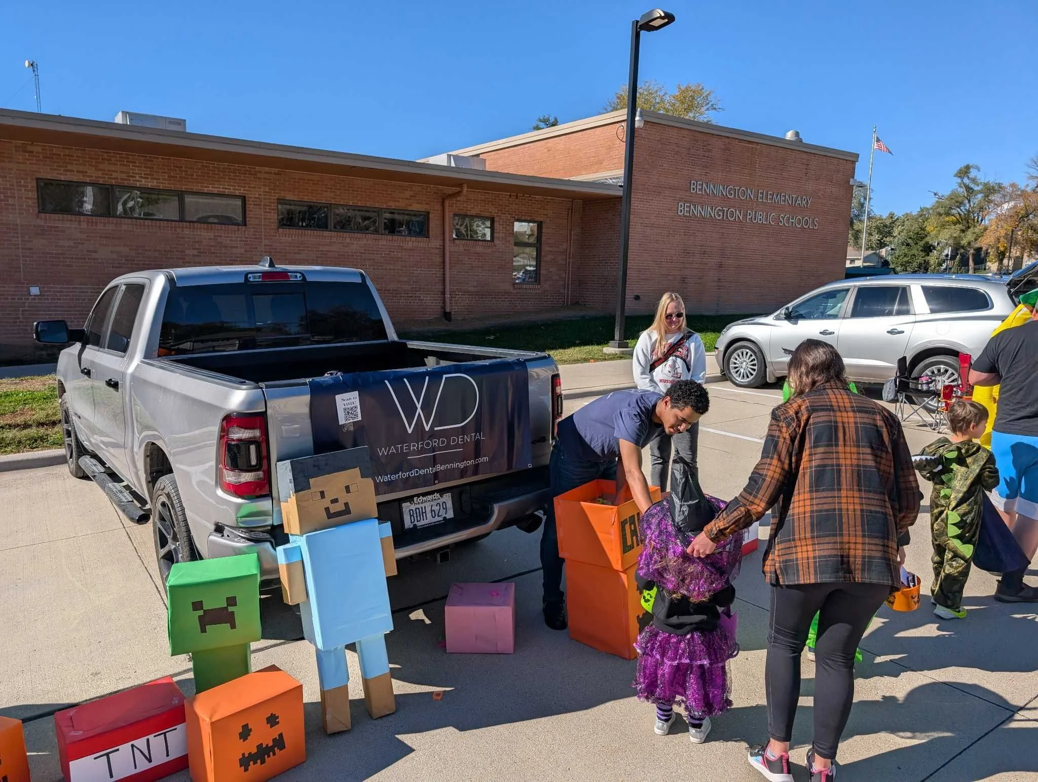 People participating in a Halloween-themed event outside Bennington Elementary School, with a colorful Minecraft-themed donation box and decorated boxes for collecting donations.