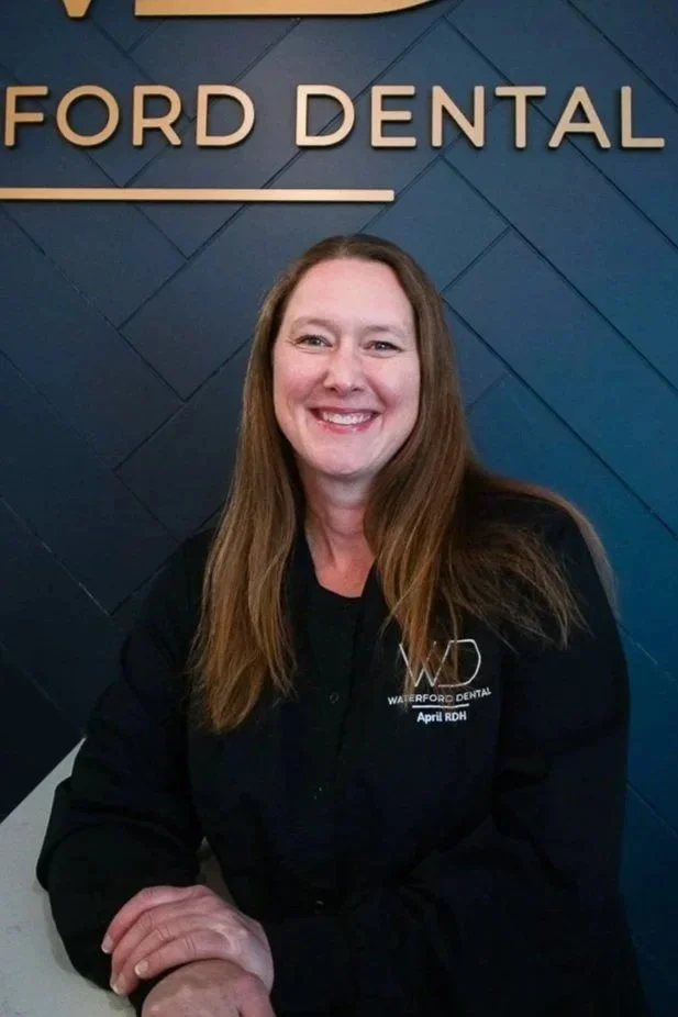 A woman with long brown hair smiling, sitting in front of a dark blue wall with gold letters spelling 'FORD DENTAL'. She is wearing a black jacket with a logo that says 'Waterford Dental' and a name tag reading 'April RDH'.