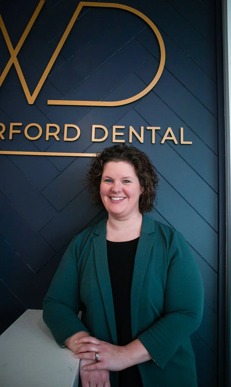 Smiling woman in teal blazer standing in front of a wall with a Dental logo at Ford Dental.