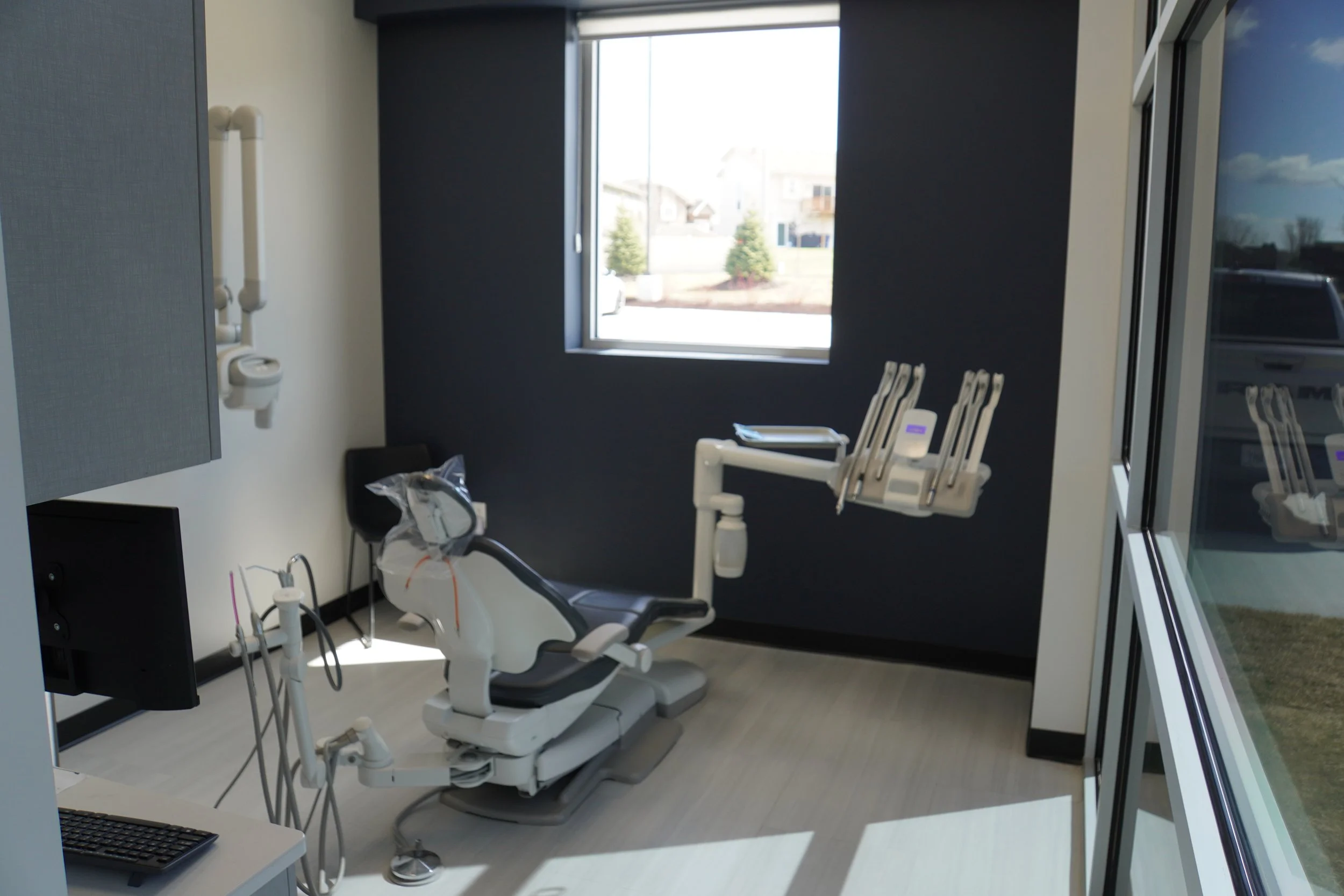 A dental treatment room featuring an adjustable dental chair, various dental instruments, and an outside view through a large window showing houses, trees, and a street.