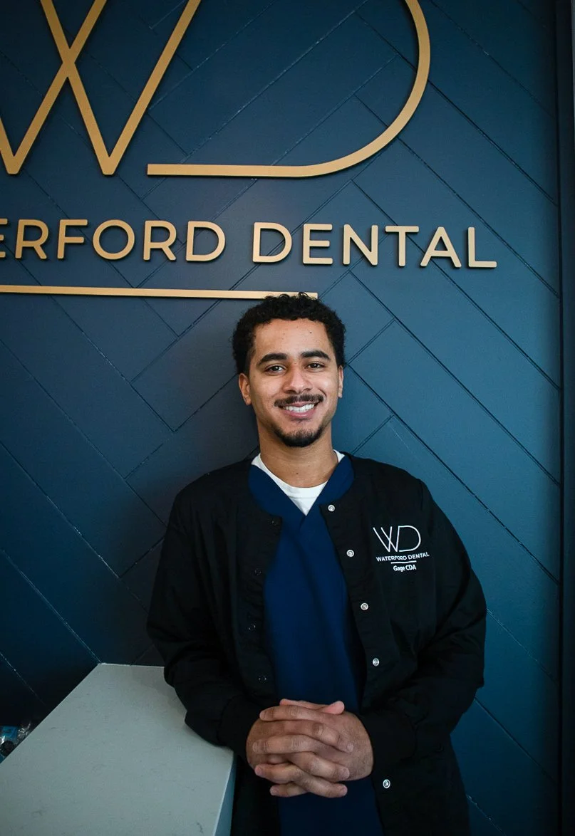 A smiling young man in a black jacket and blue scrubs standing in front of a blue wall with the words "Waterford Dental" and a logo.