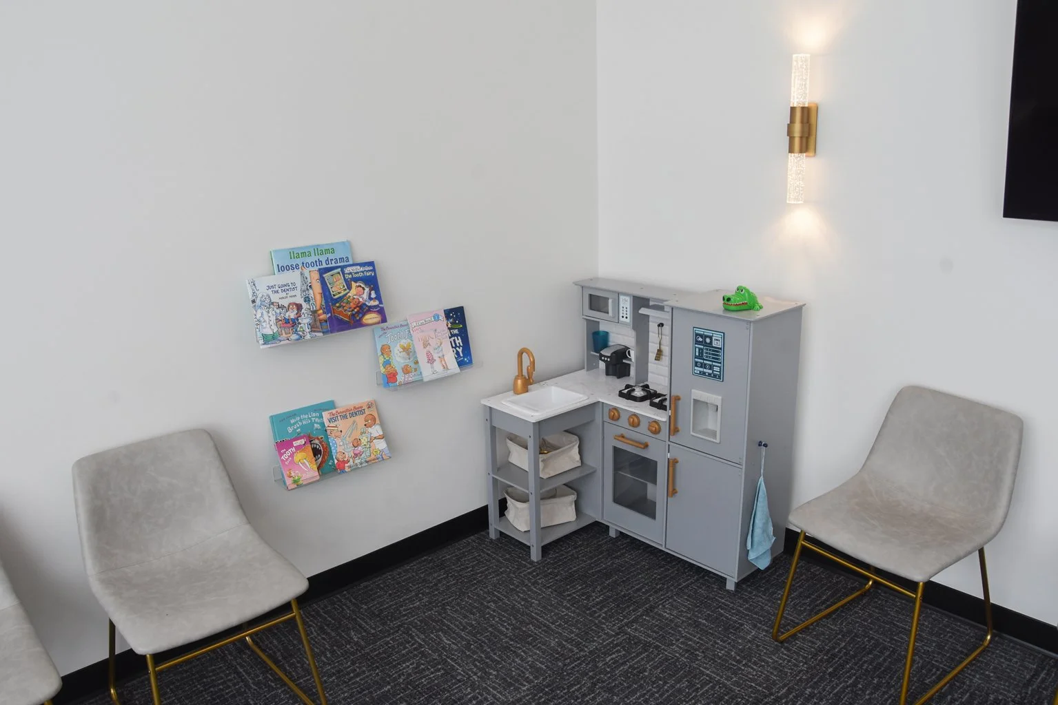 Children's play area in a dental office with a small pretend kitchen set, two white chairs with gold legs, and books about dental health on wall shelves.
