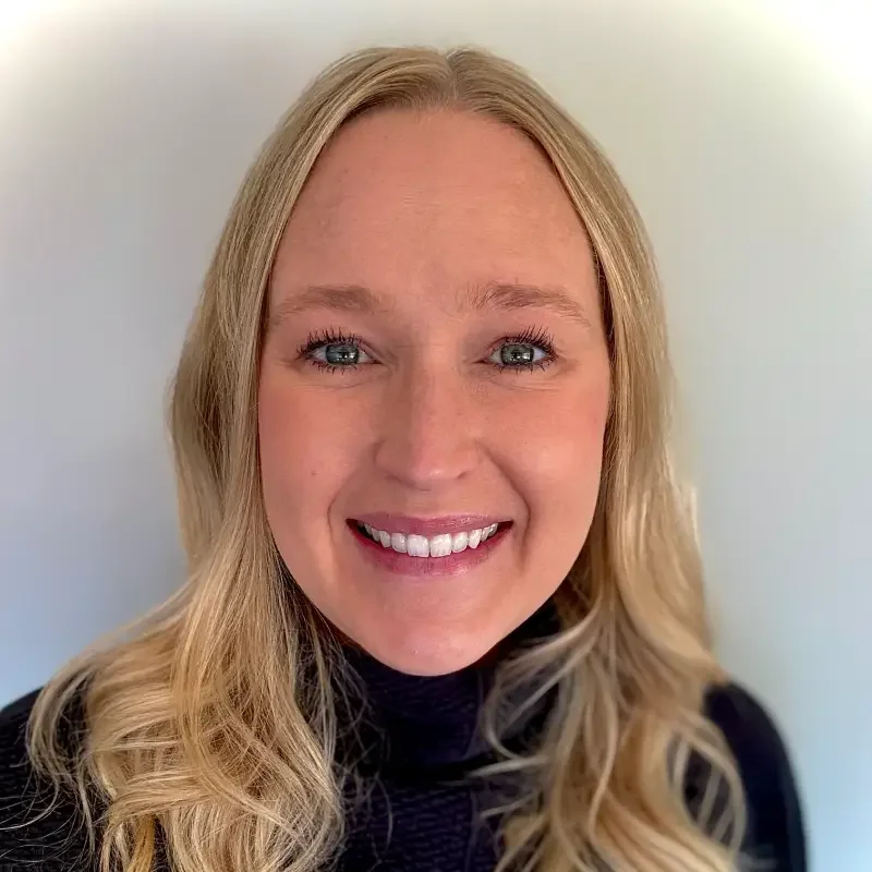 Close-up of a blonde woman smiling, wearing a black top, against a plain light-colored background.