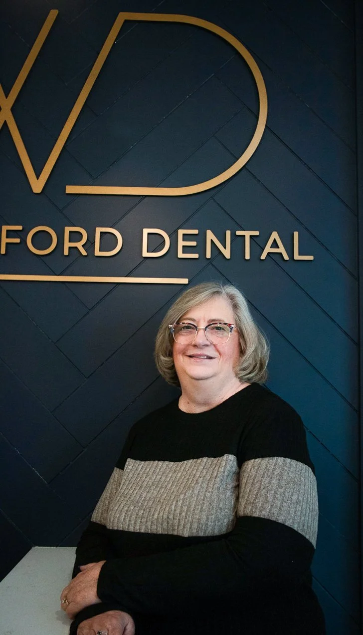 A woman with gray hair and glasses standing in front of a wall with the logo and name of Ford Dental.