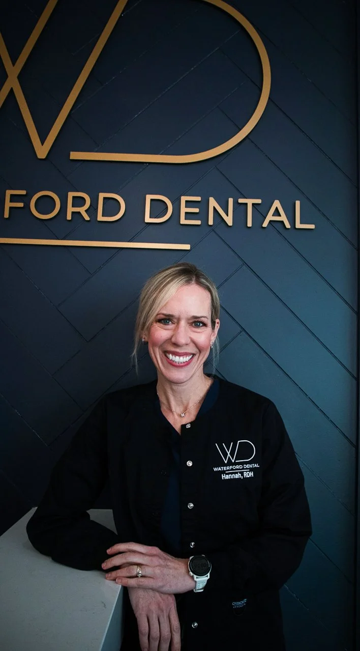 A smiling woman in a black dental uniform with "Waterford Dental Hannah, RDH" embroidered on it, standing in front of a blue wall with the 'Ford Dental' sign in gold letters.
