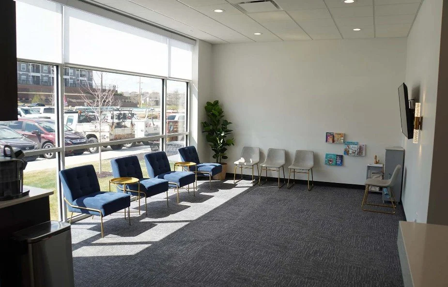 An empty waiting room with blue and beige chairs, a small bookshelf with children's books, a wall-mounted TV, a tall green plant, and large windows showing parked cars outside.