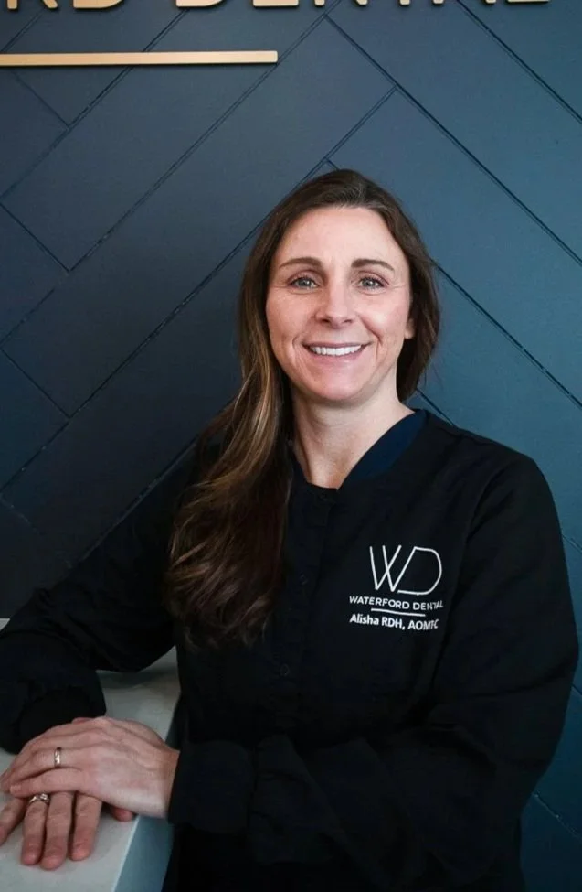 A woman with long brown hair smiling at the camera, wearing a black lab coat with the logo of Waterford Dental and her name, Alisha RDH, on it, sitting at a white surface against a dark blue wall.