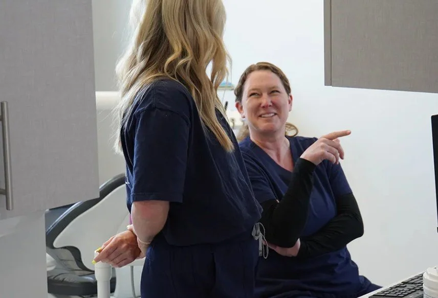 Two healthcare workers in navy scrubs talking and smiling in a medical office, one pointing at a computer screen.