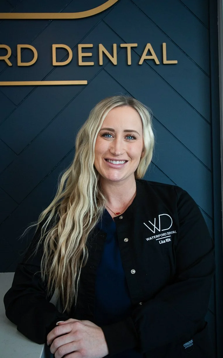 A woman with long blonde hair smiling, wearing a black jacket with the logo 'Waterford Dental' and the name 'Liza Roh,' sitting in front of a blue wall with the words 'Waterford Dental' partially visible.