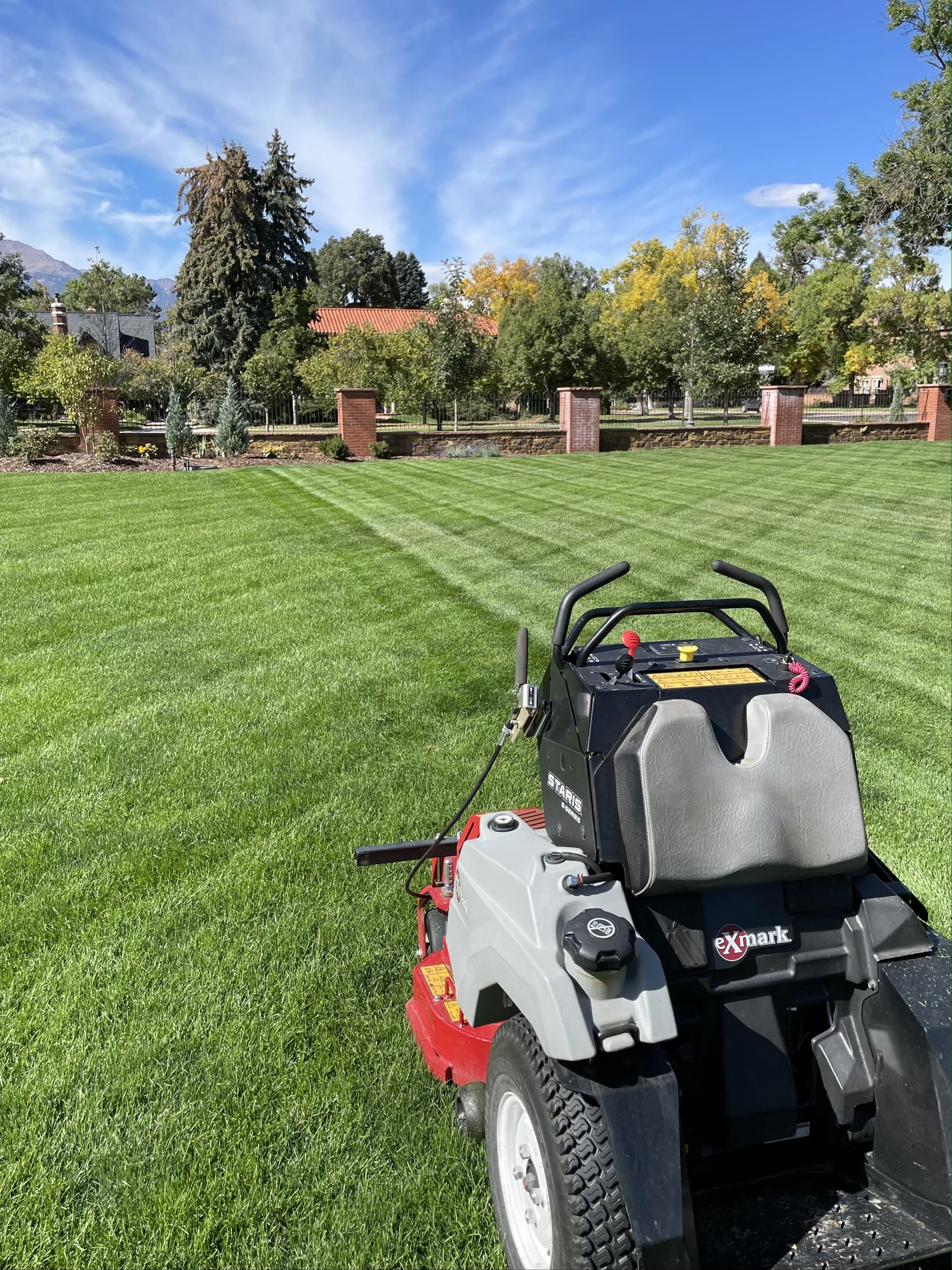 Lawn mower on a freshly cut green lawn with a park and trees in the background under a partly cloudy sky.