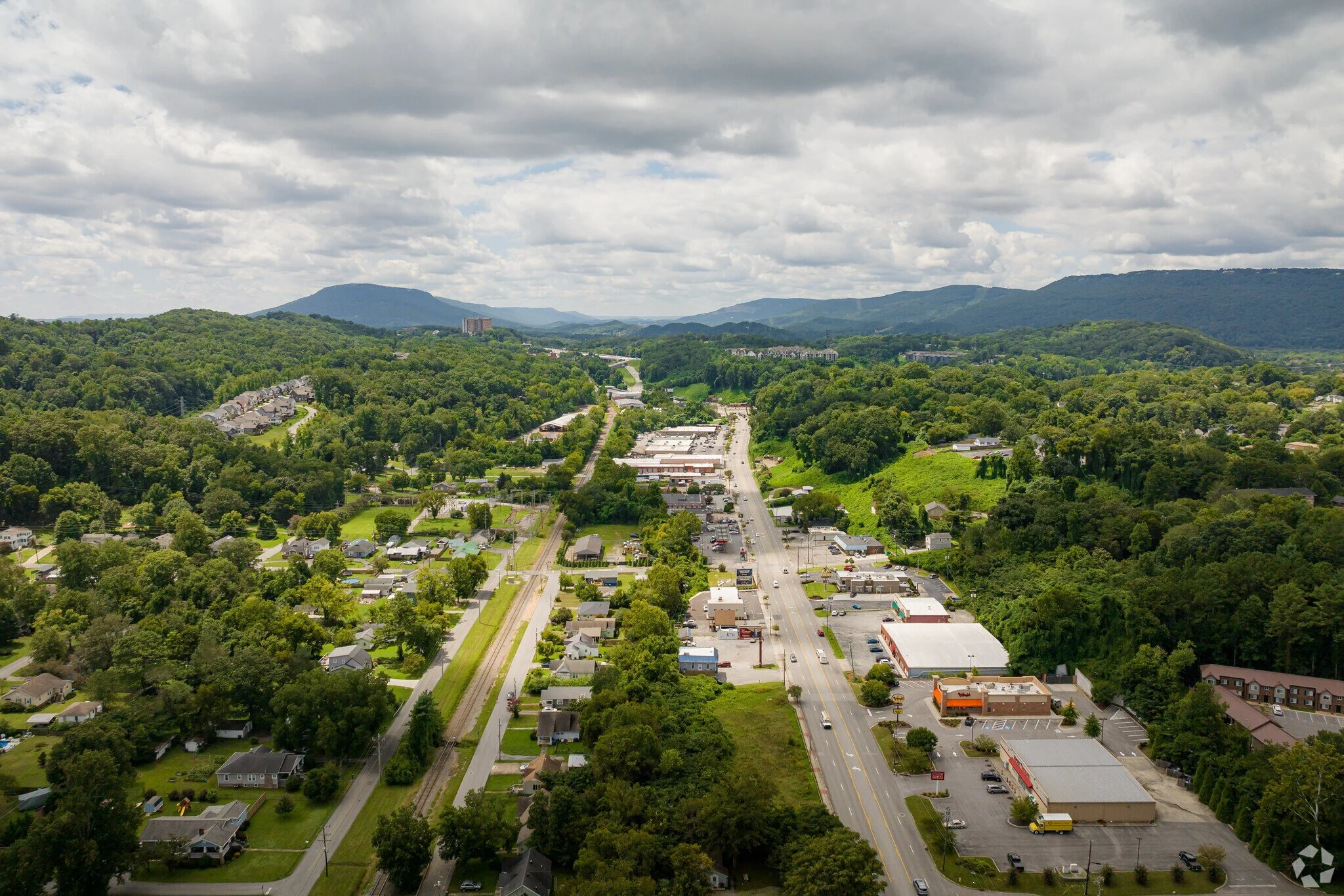Aerial view of a small town with a main road running through it, surrounded by lush green trees and hills, with mountains in the background under a partly cloudy sky.