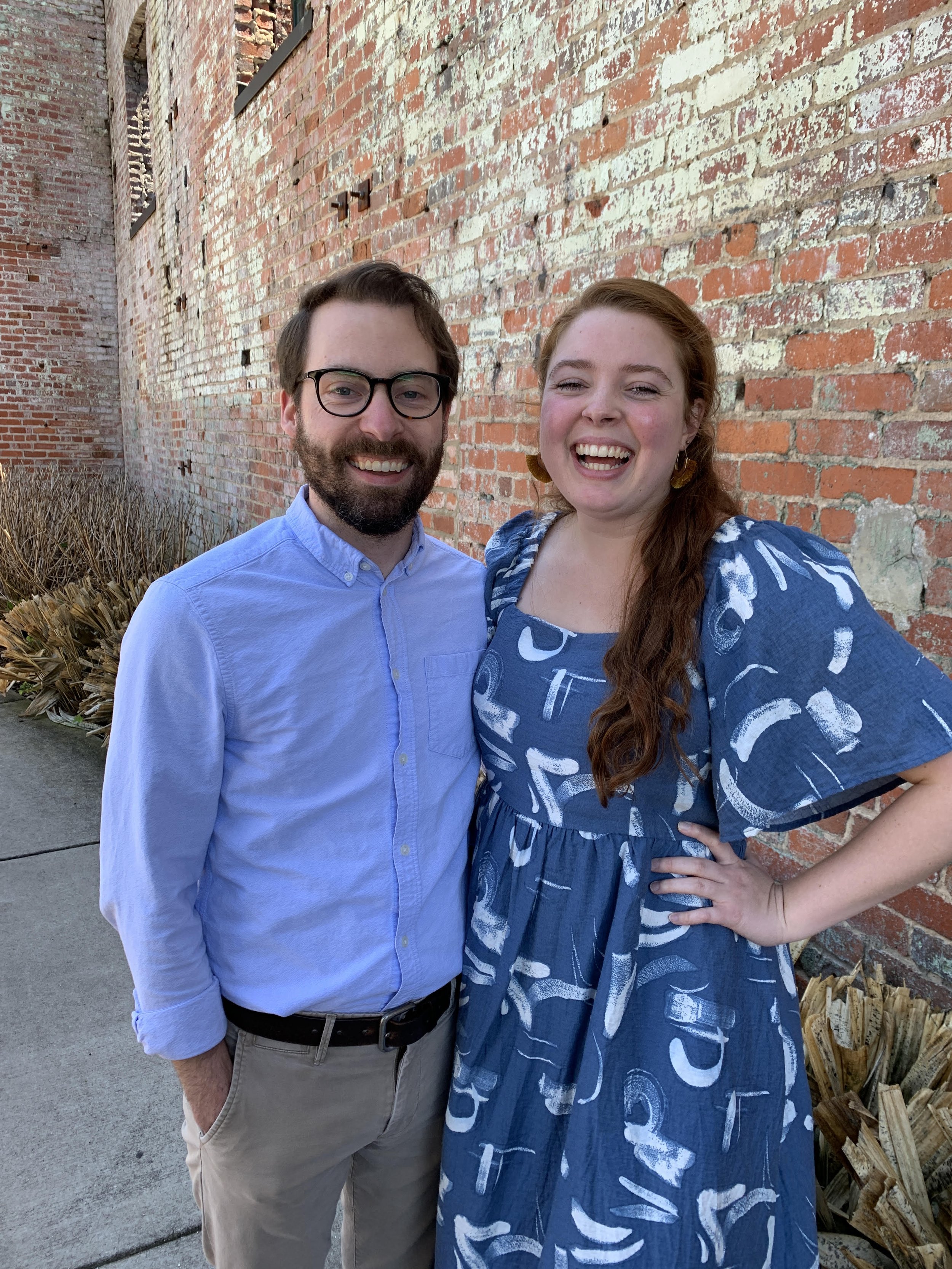 A smiling man with glasses and a beard in a light blue shirt standing next to a smiling woman with red hair in a blue patterned dress, standing beside a brick wall.
