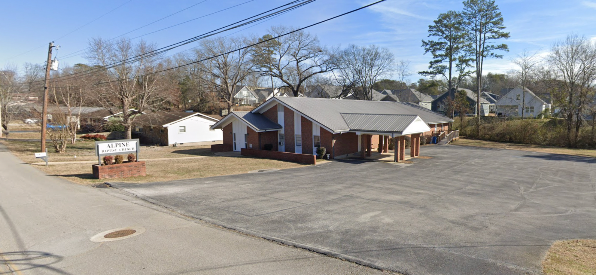 Exterior view of Alpine Baptist Church with a brick facade, parking lot, and surrounding trees in a suburban neighborhood.