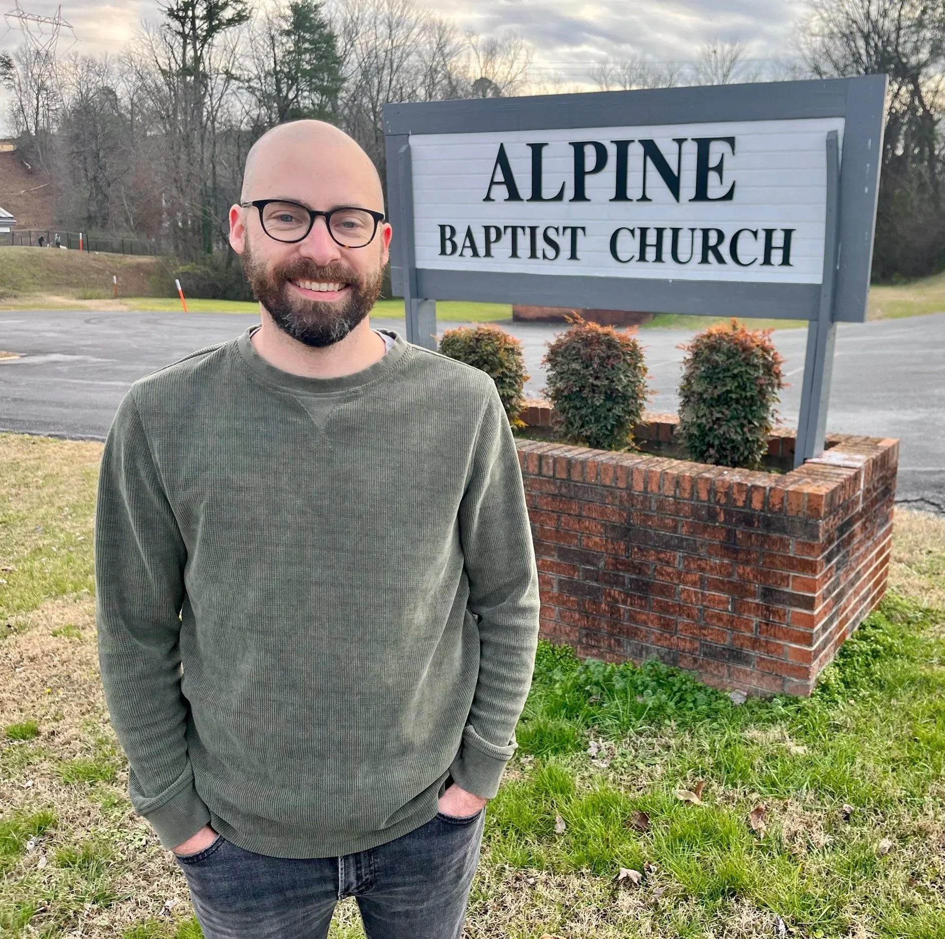 A man with glasses and a beard standing outdoors on grass next to a sign that reads 'Alpine Baptist Church'. The background features trees and a parking lot.