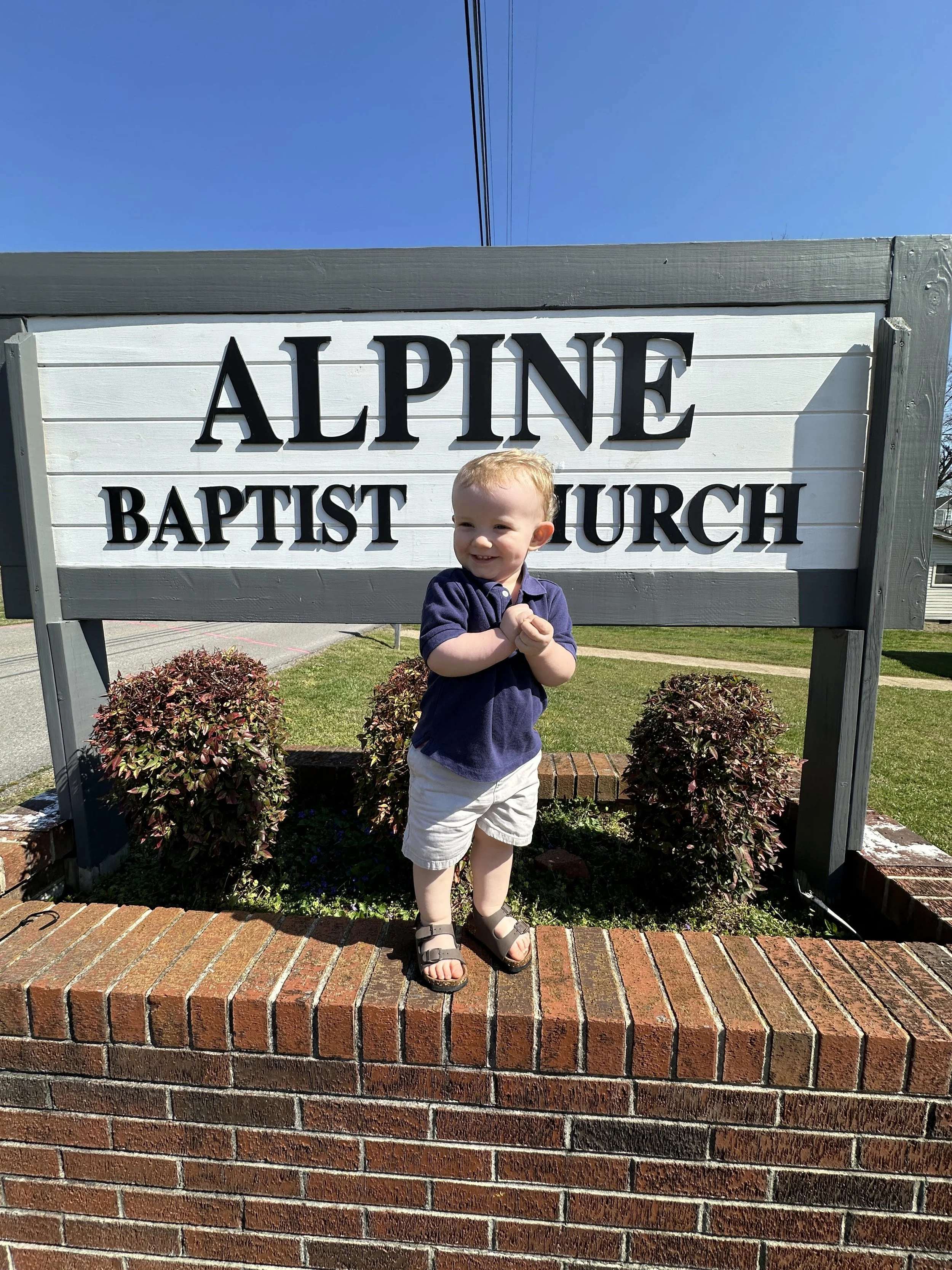 A young boy with blonde hair, wearing a navy blue shirt, khaki shorts, and sandals, standing on a brick ledge in front of a sign that reads 'ALPINE BAPTIST CHURCH' on a sunny day with a clear blue sky.