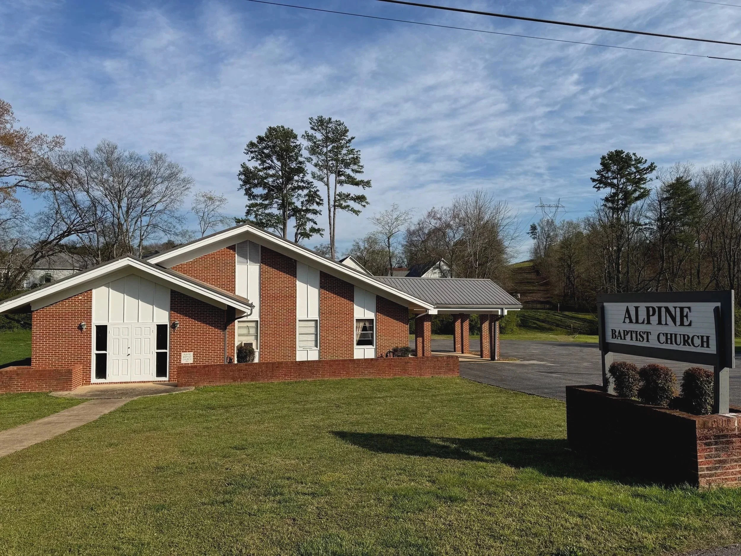 Exterior view of Alpine Baptist Church, a brick building with white accents, surrounded by a well-maintained lawn, and a sign with the church's name near the road.