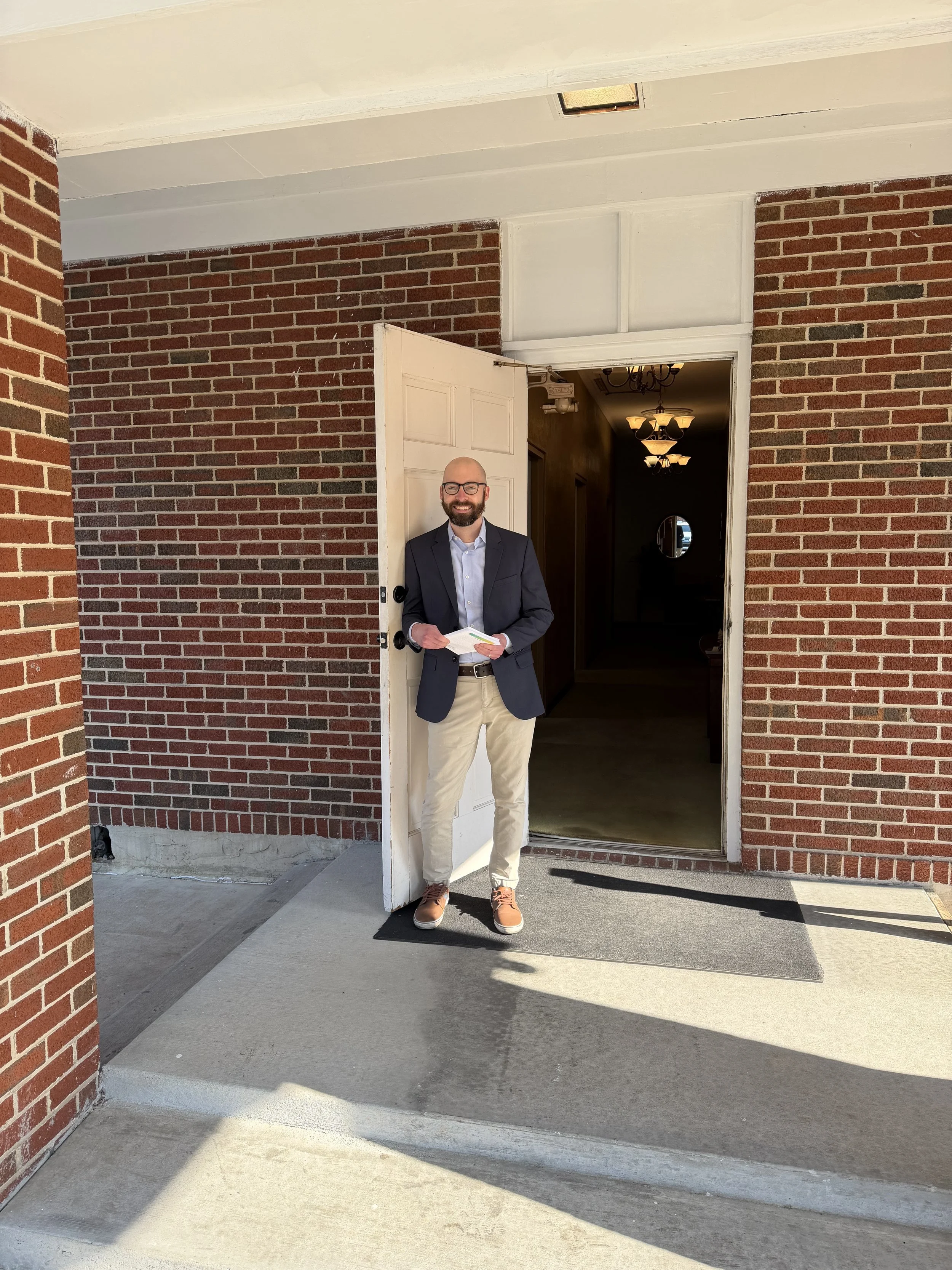Man standing at a house entrance, holding papers, smiling, dressed in a blazer, shirt, khaki pants, and brown sneakers, with a brick wall and interior lights visible behind him.