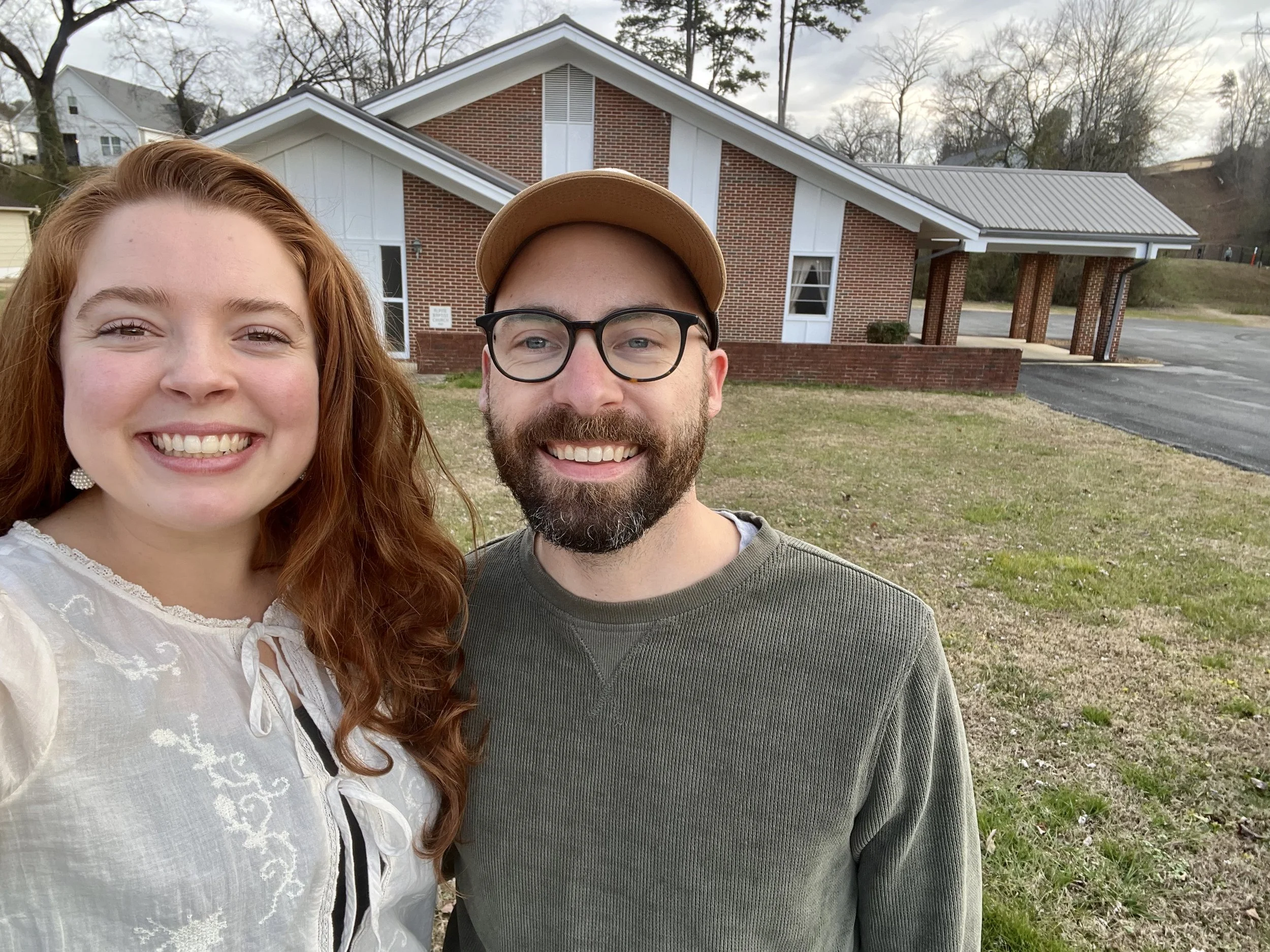 A smiling woman with red hair and a man with glasses, a beard, and a tan cap, standing outside a brick building during daytime.