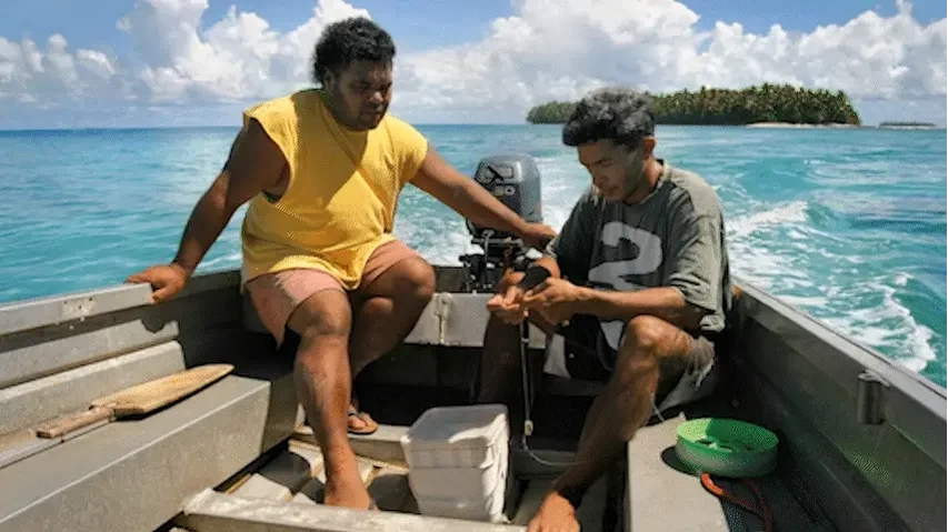 Two men on a small boat in the Pacific ocean, with a small island in the background.