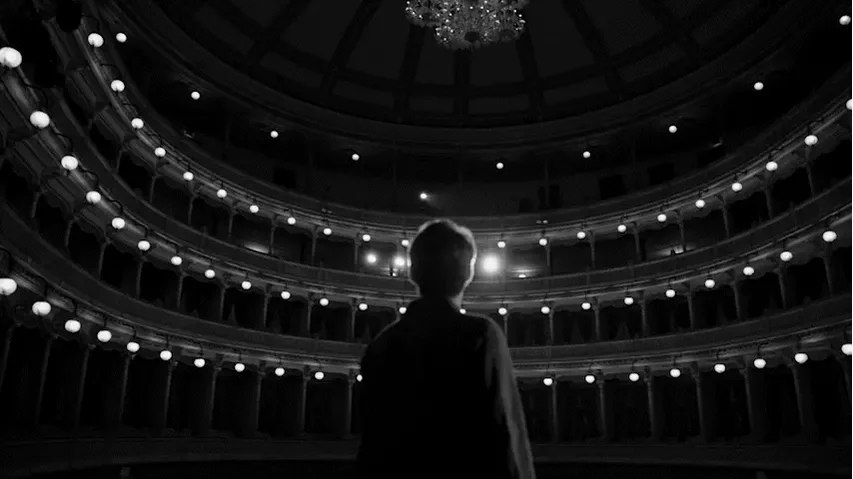 A person standing in an empty theater, facing the stage, with ornate balcony seats and a chandelier hanging from the ceiling.