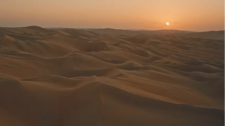Desert landscape with sand dunes at sunset, orange sky with the sun near the horizon.