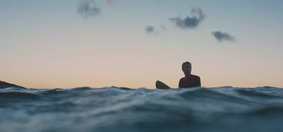 A person holding a surfboard sitting on a surfboard in the ocean during sunset, with a few clouds in the sky.