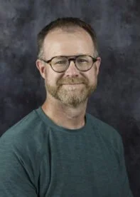 Portrait of a man with glasses, short hair, and a beard, wearing a green shirt, smiling against a dark textured background.