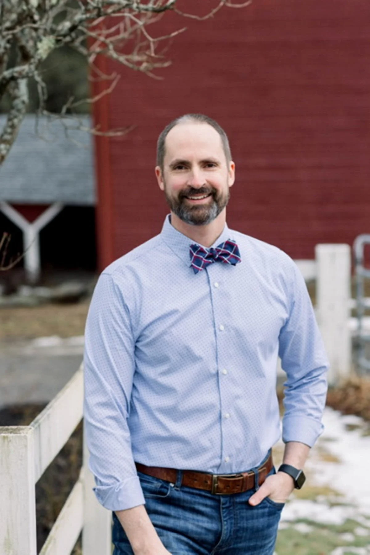 A man with a beard and mustache wearing a blue dress shirt, a red and blue plaid bowtie, jeans, and a watch, standing outdoors near a white fence, with a red building and leafless trees in the background.