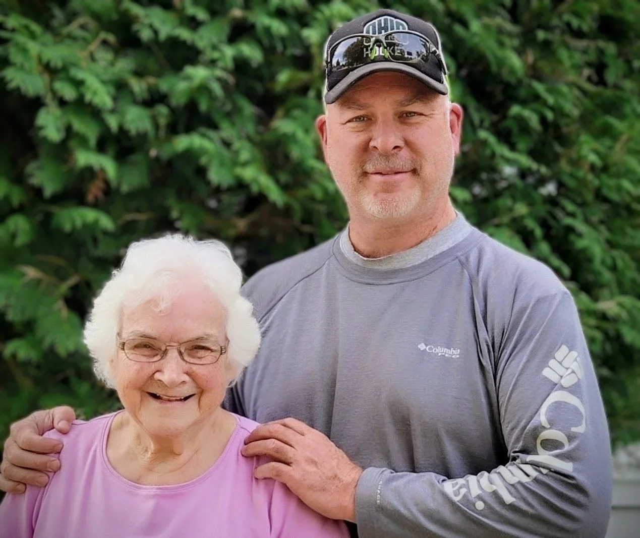 A smiling elderly woman with white hair and glasses, standing next to a smiling middle-aged man wearing a gray long sleeve shirt and a cap, outdoors with green bushes in the background.