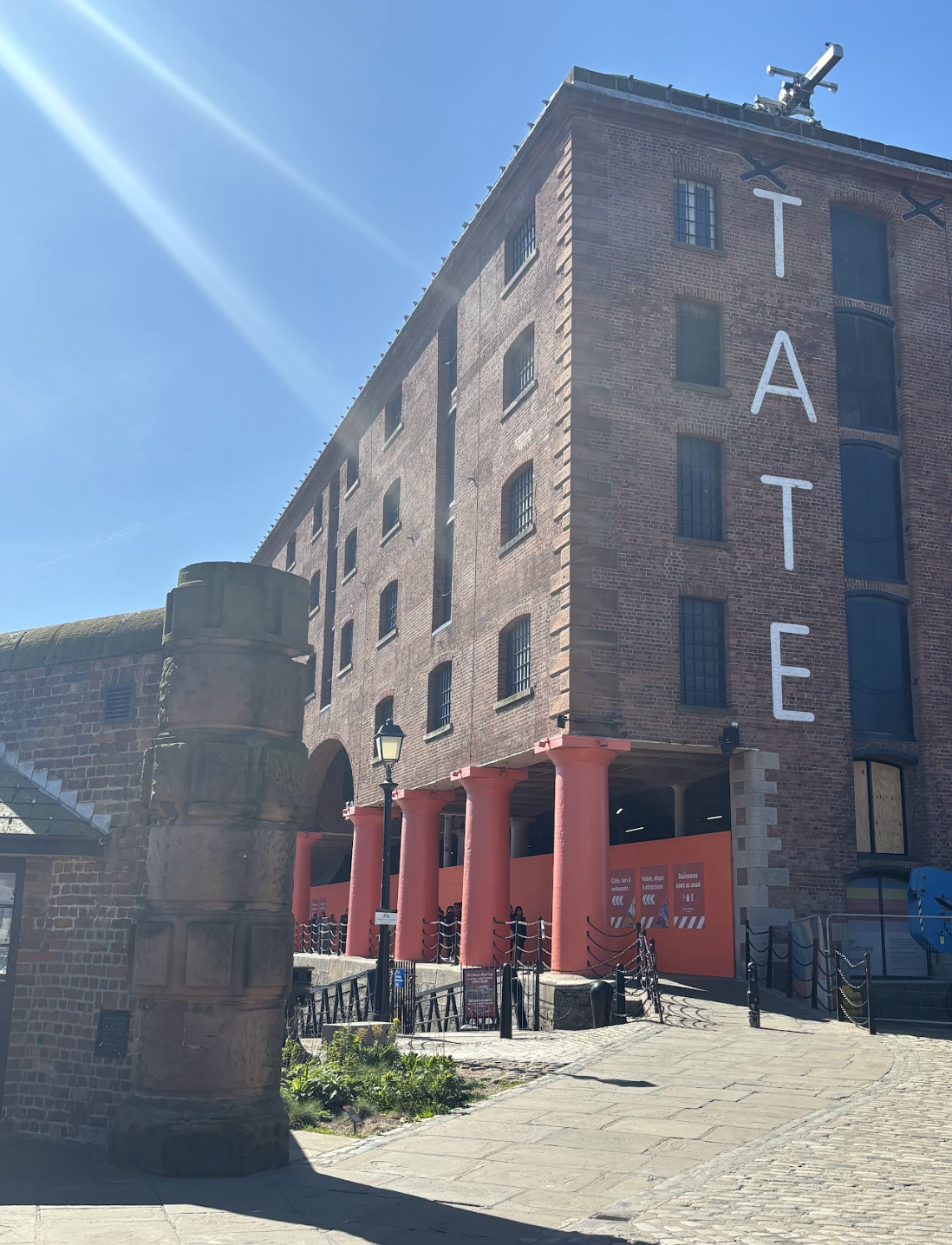 A brick building with the large white letters spelling 'TATE' vertically on the side, with an open ground floor supported by red columns, a lamppost, and a cobblestone sidewalk.