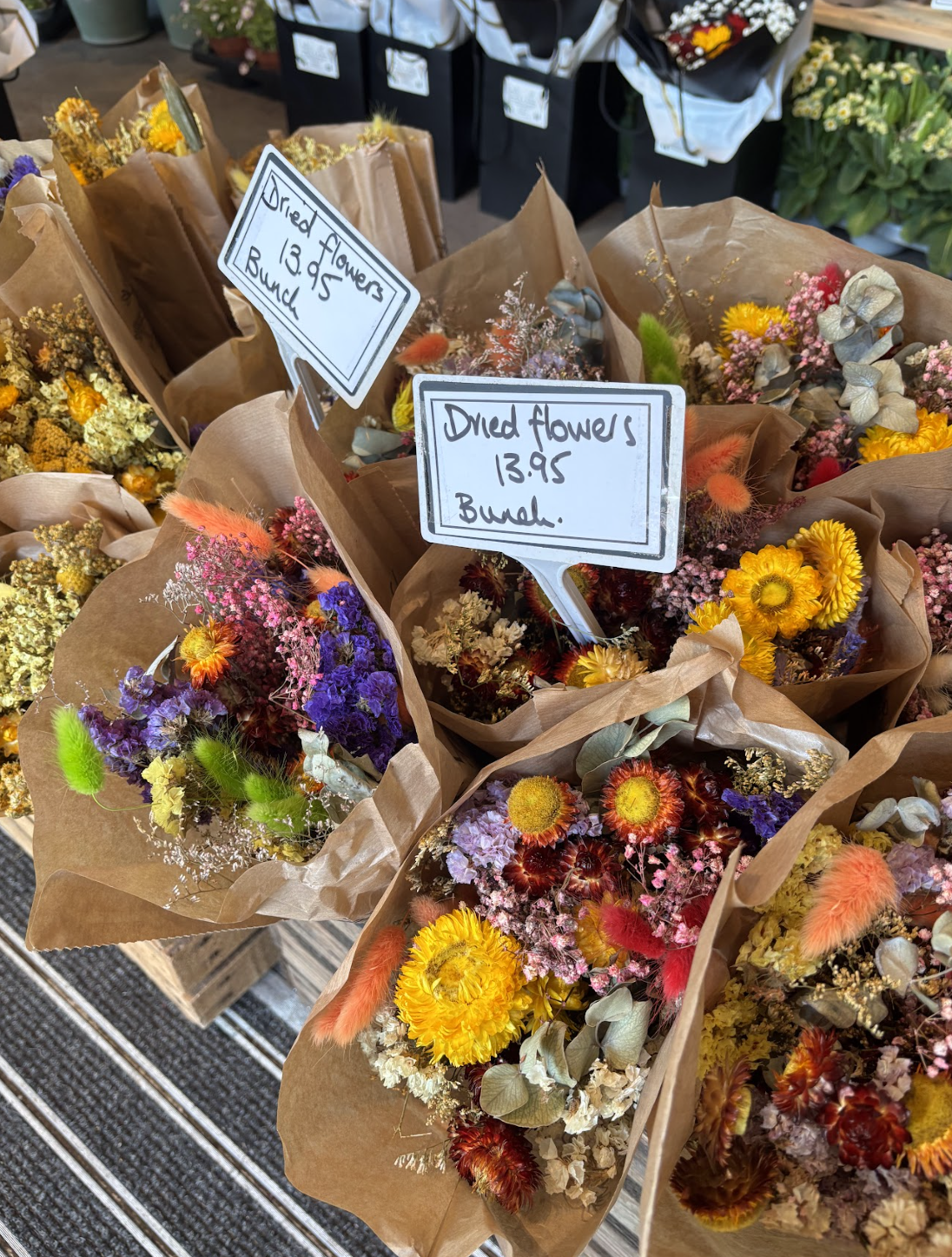 Multiple bouquets of dried flowers wrapped in brown paper on display at a store, with handwritten signs indicating prices and types of flowers, including one sign reading 'Dried flowers 13.95 a bunch'.