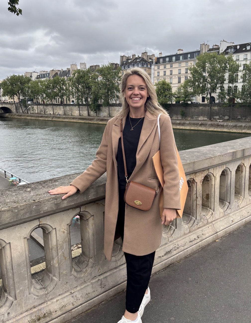 A smiling woman in a beige coat and black pants standing on a bridge over a river in Paris, France, with classic Parisian buildings and trees in the background.