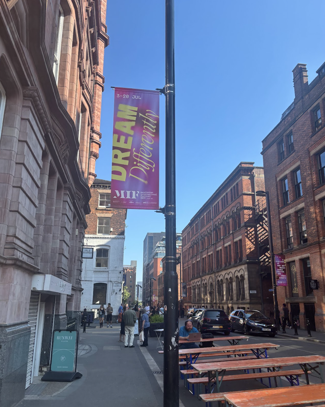 Urban street scene with buildings, people, parked cars, and outdoor tables under a bright blue sky. A banner on a street lamp promotes the 'DREAM Differently' event from July 3 to July 20.